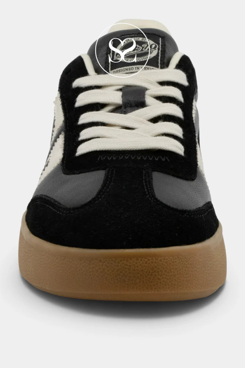 A close-up front view of a low-top sneaker featuring black suede and dark gray panels, off-white laces, and a gum brown sole.