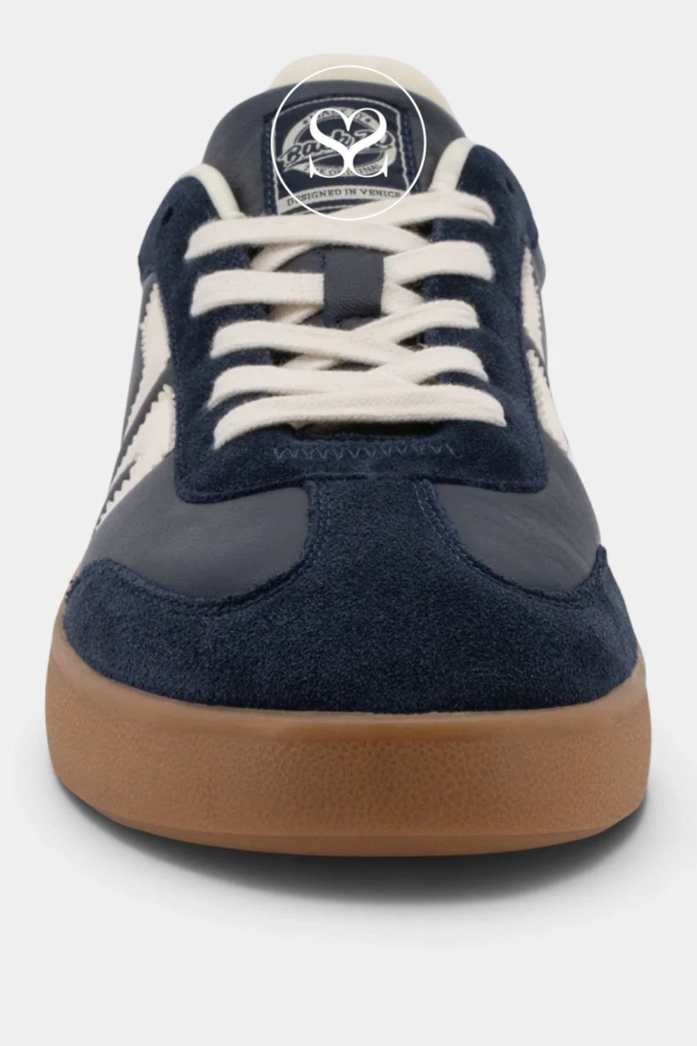 A close-up front view of a low-top sneaker featuring navy blue suede and smooth leather panels, off-white laces, a cream tongue label with a black heart logo, and a brown gum rubber sole, set against a light gray background.