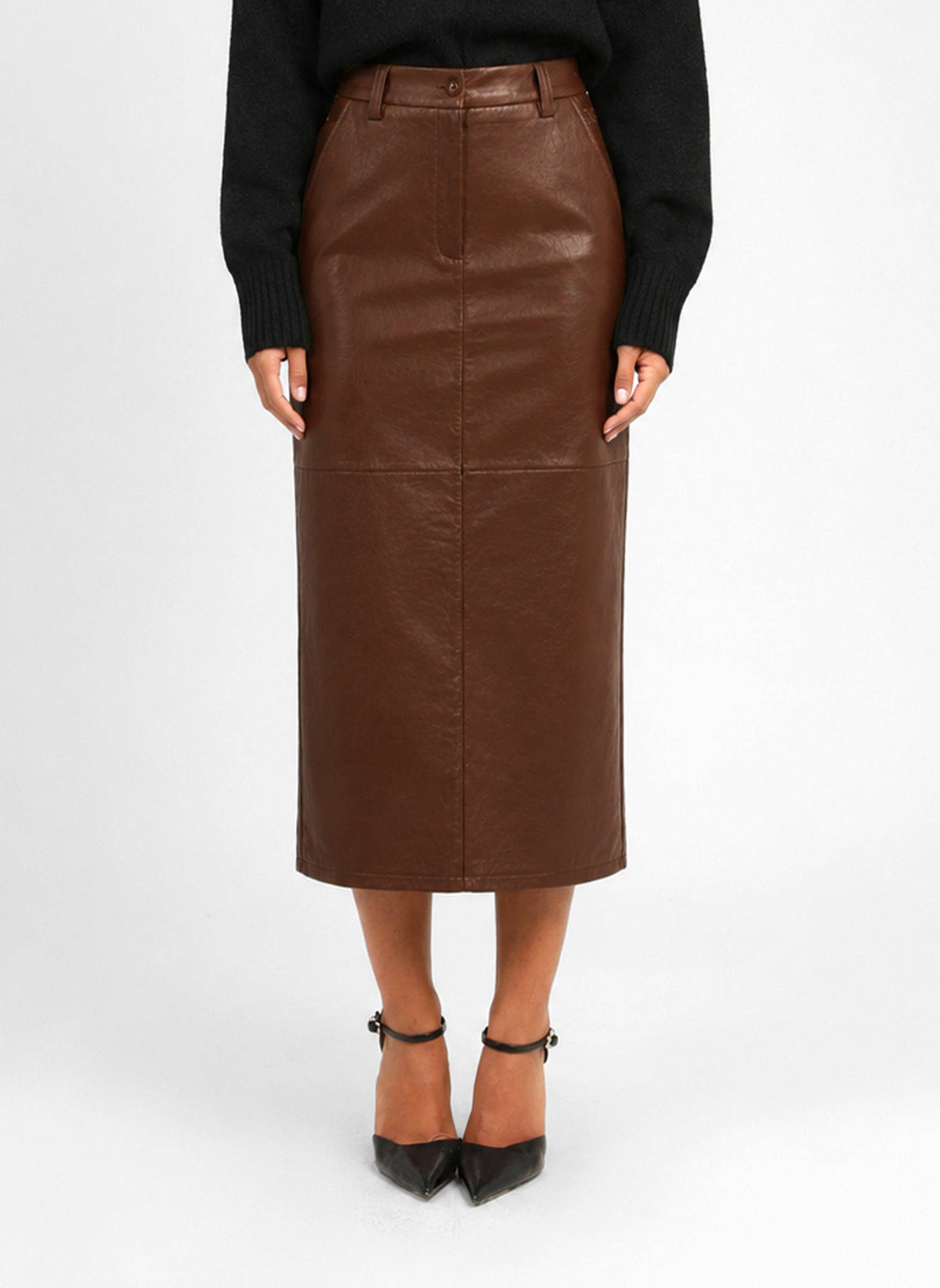 A model wearing a brown faux-leather midi skirt with a button waistband, paired with a black sweater and black pointed-toe heels, shown against a plain white studio background.