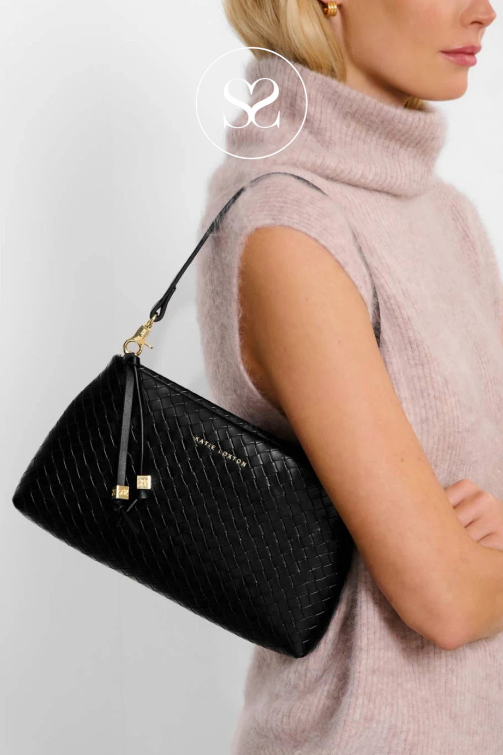 A woman wearing a light pink sleeveless turtleneck sweater carries a black woven leather shoulder bag with gold hardware and a small gold logo on the front, against a light gray background.