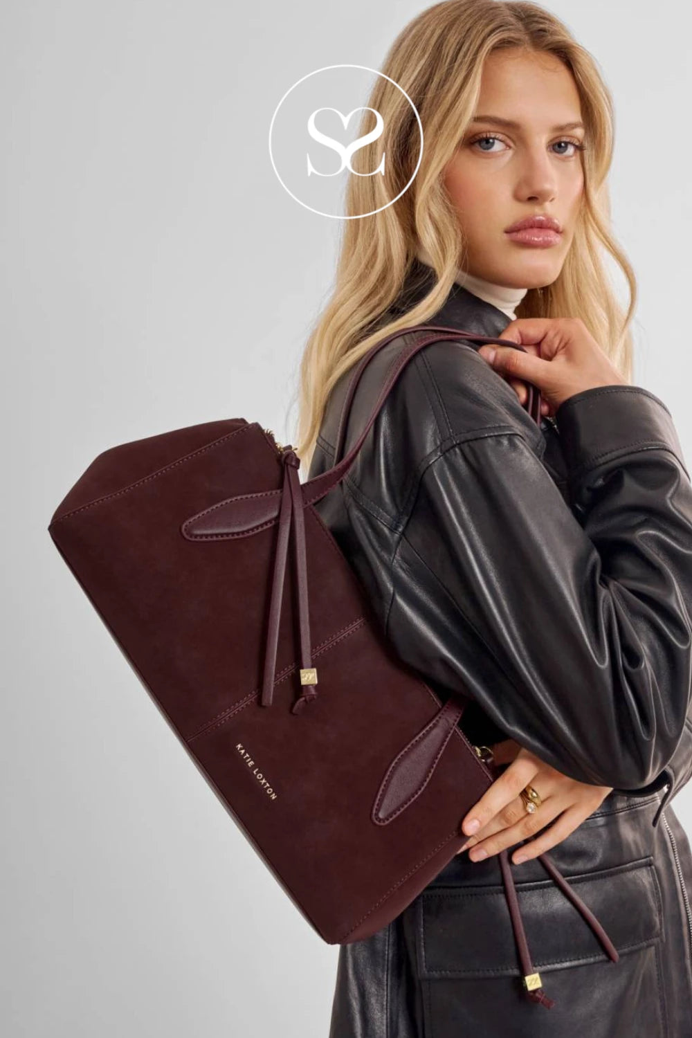 A woman in a black leather jacket carries a large burgundy suede tote bag with a drawstring and gold-tone hardware against a light gray studio background