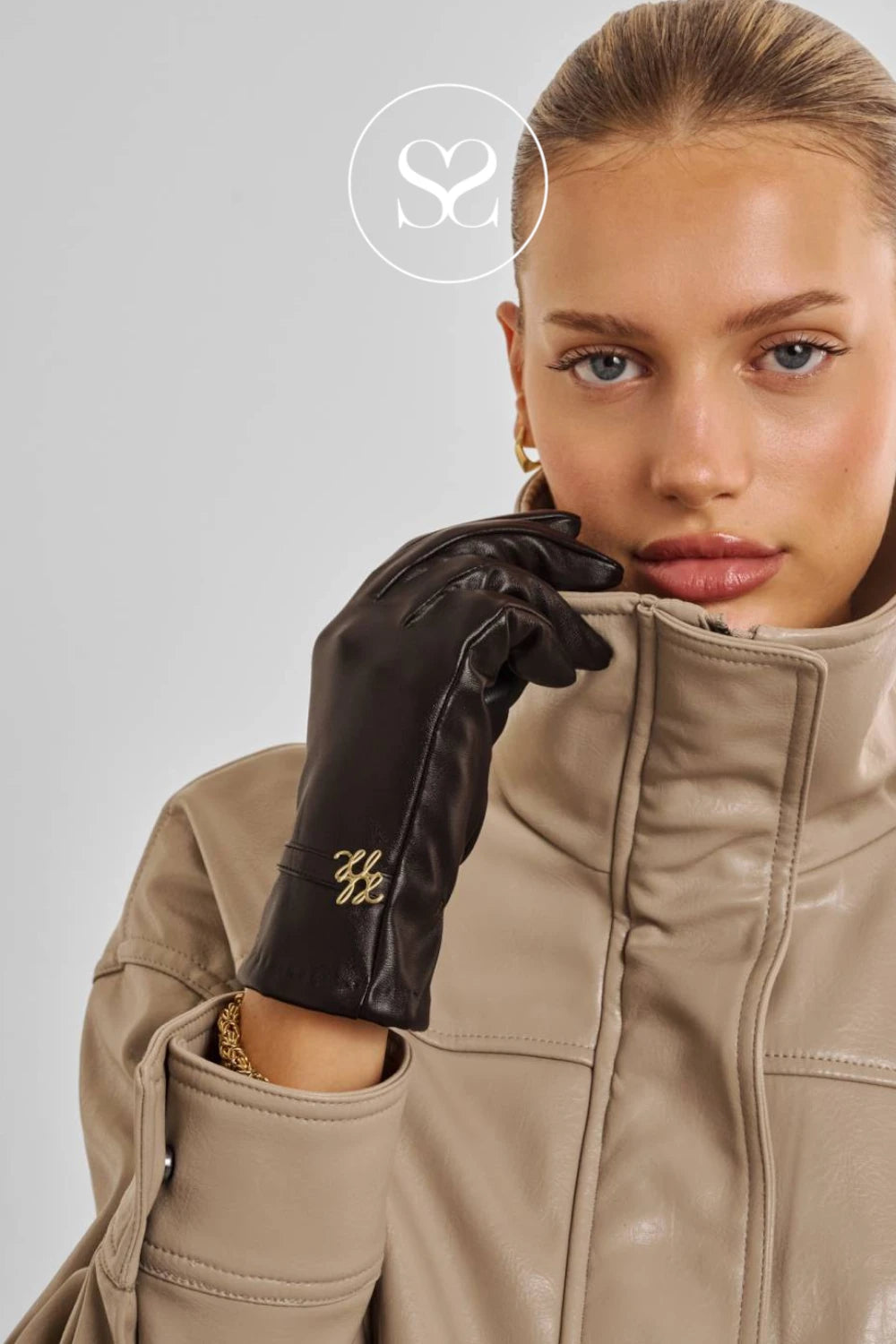 A close-up of a model wearing a black leather glove with a gold embroidered monogram logo, paired with a beige faux-leather jacket. A white circular logo overlay with 'S2' is visible at the top center.