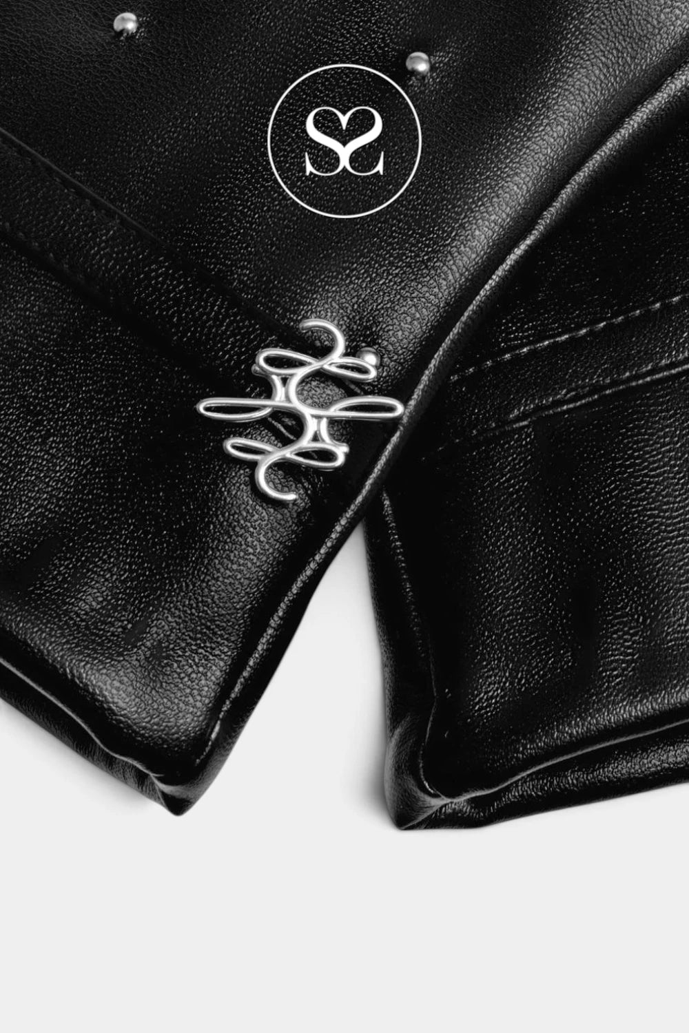 Close-up of a black pebbled leather garment, likely a jacket or vest, featuring silver-tone metal studs, a silver interlocking logo charm, and a white circular logo overlay on a light gray background.