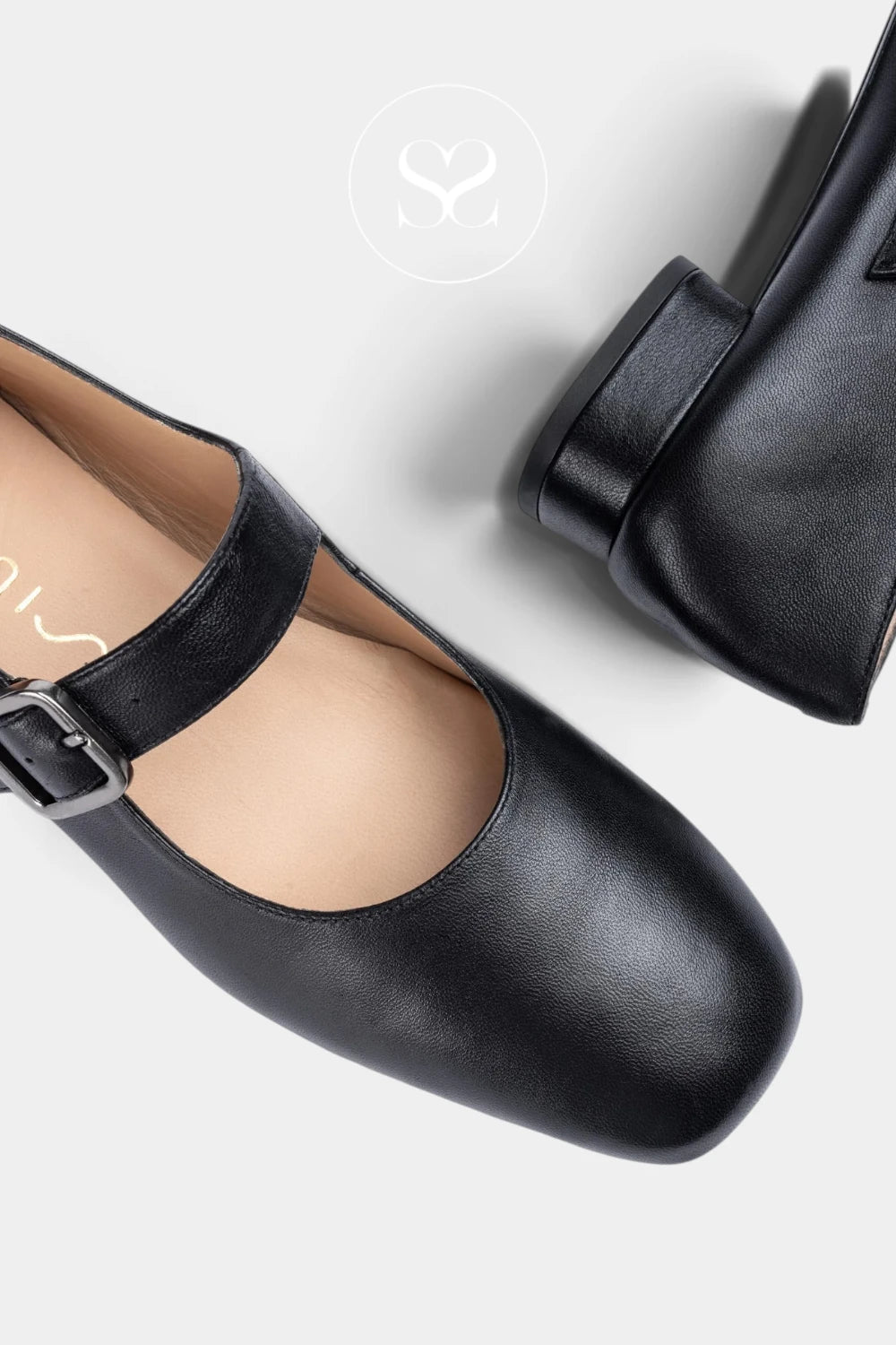 A pair of uNISA black Mary Jane flats with square toes and adjustable buckle straps, shown on a light gray background with a faint brand logo watermark.