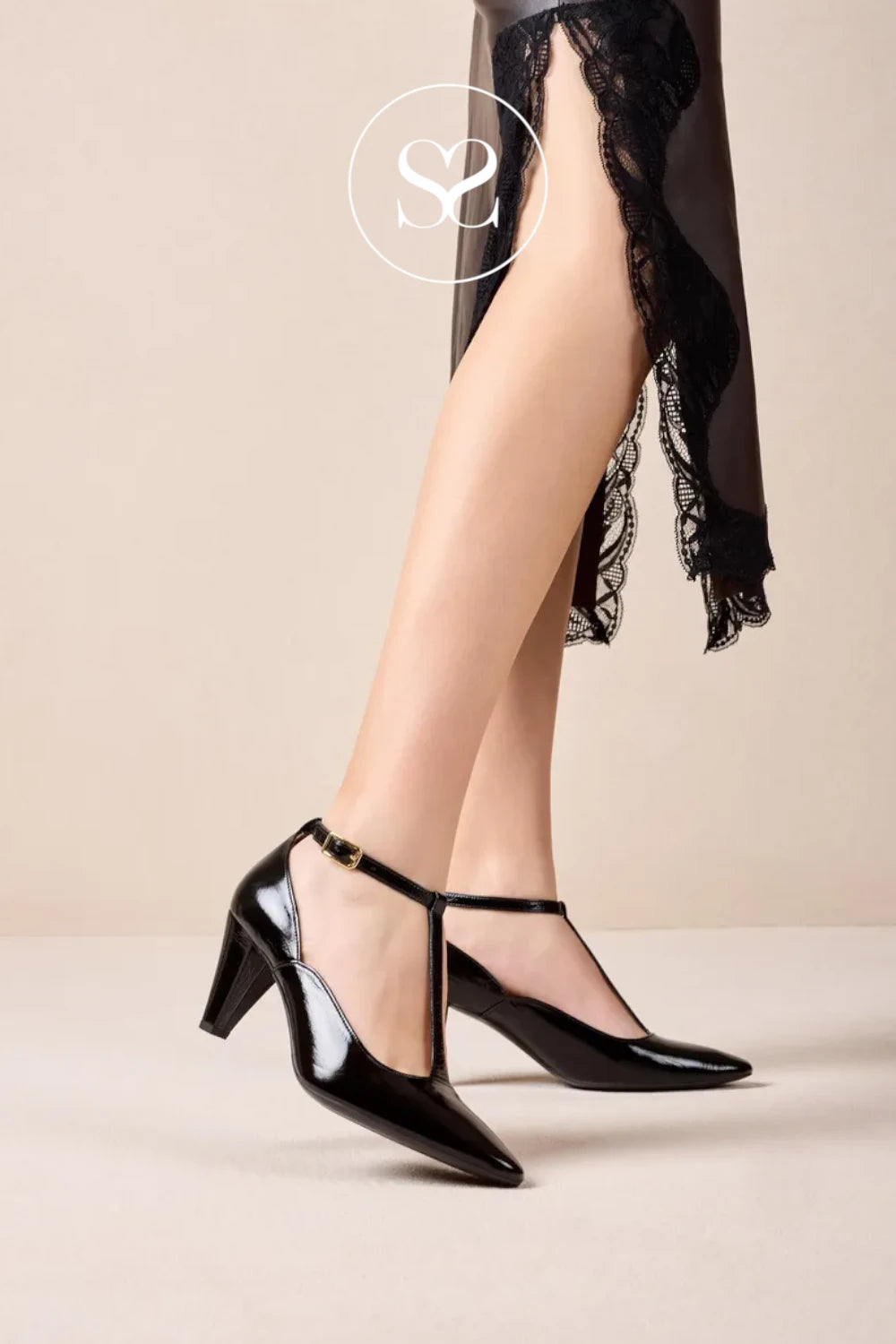 A pair of black patent leather T-strap high heels with kitten heels, shown on a model's feet against a neutral beige background. The model is wearing sheer black lace-trimmed trousers, and a circular 'S2' logo watermark is visible at the top.