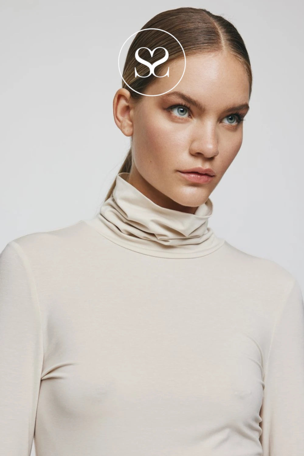A close-up of a woman wearing a beige long-sleeve turtleneck top, with a white circular logo overlay on her forehead, set against a plain light gray background.