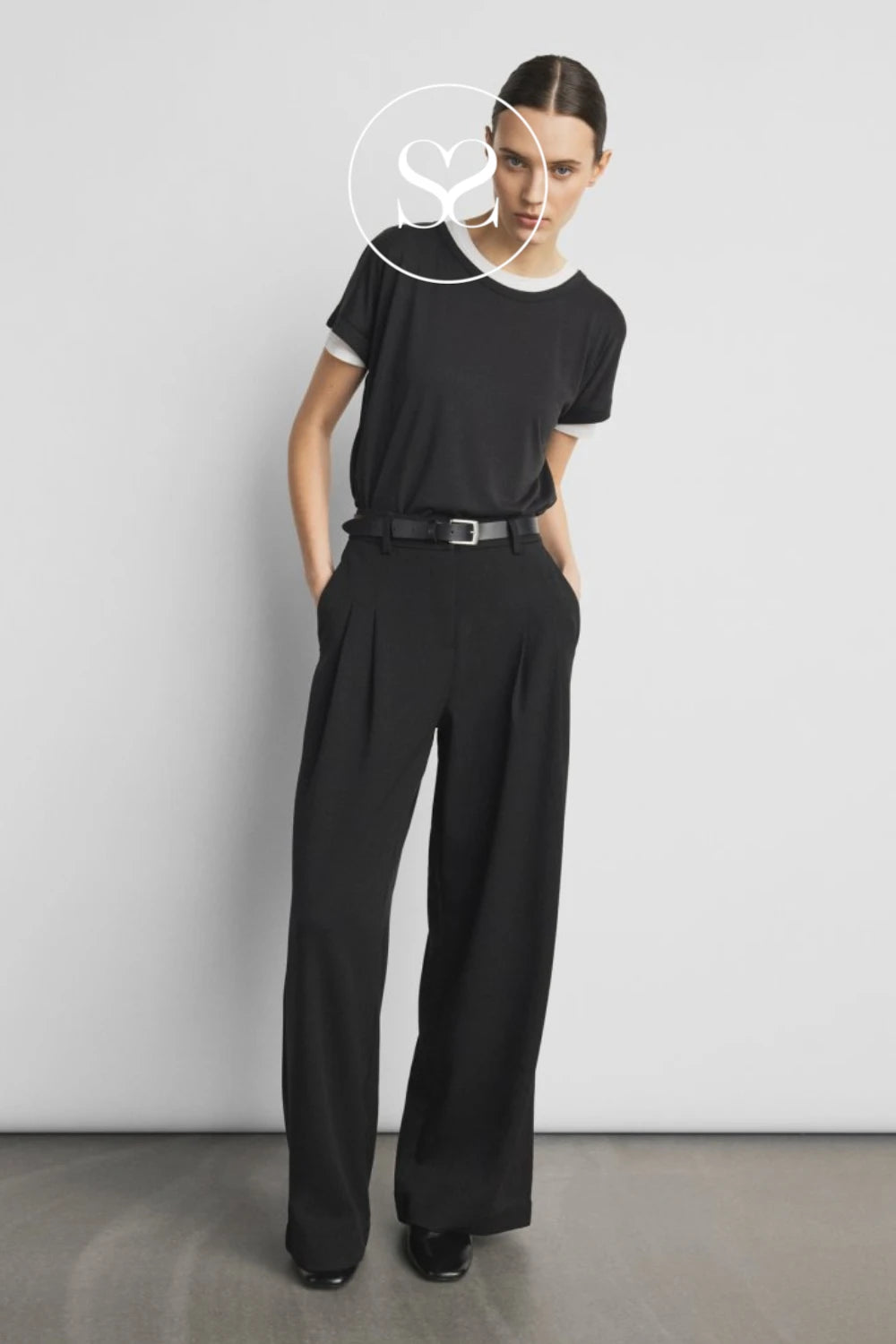 A model stands against a light gray studio wall wearing a black short-sleeve top with white trim and black wide-leg trousers, styled with a black belt. A white circular logo overlay is visible near the neck.