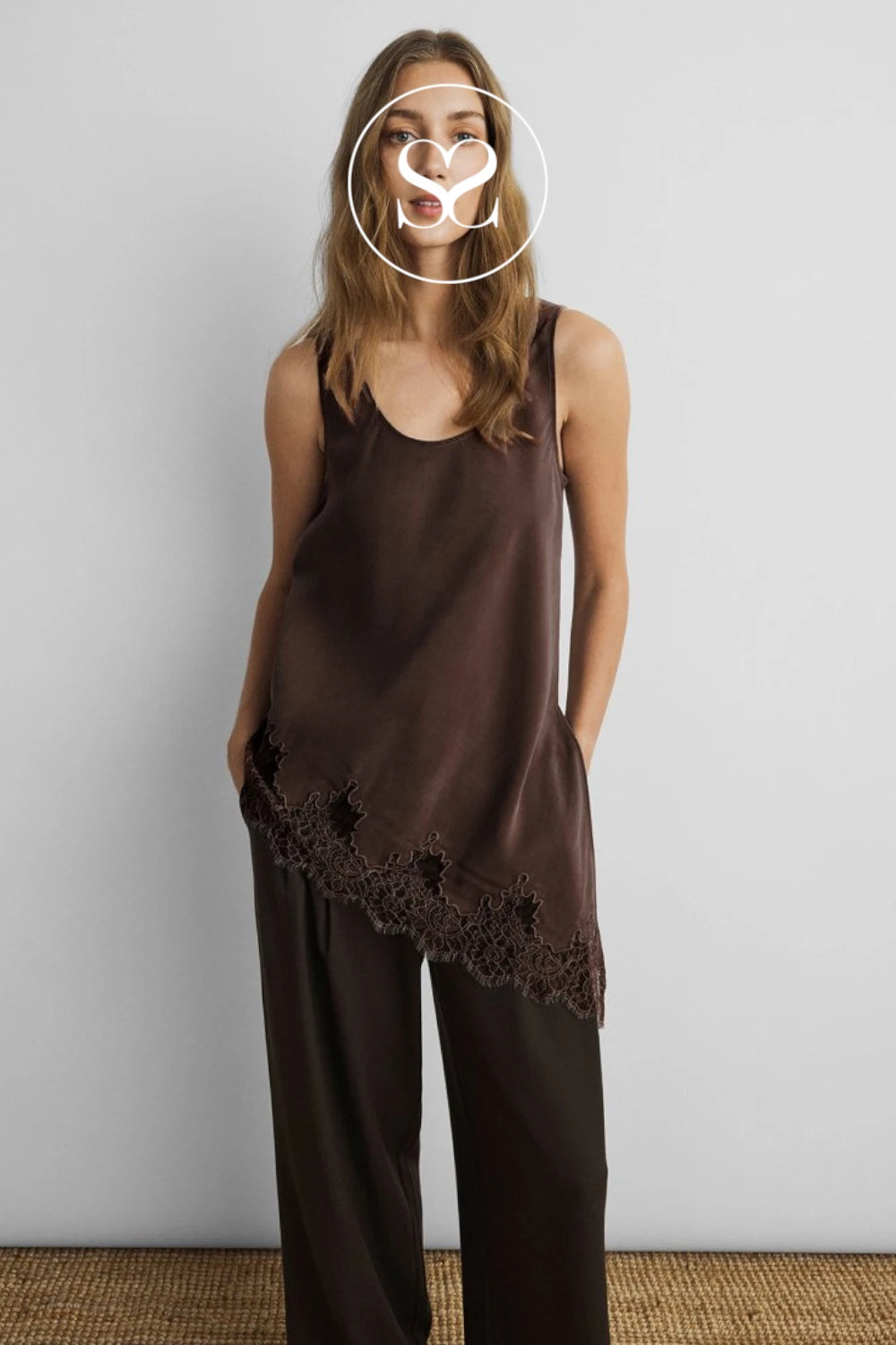 A woman modeling a dark brown satin-like sleeveless top with an asymmetrical lace-trim hem, paired with matching wide-leg pants, against a plain light gray wall.