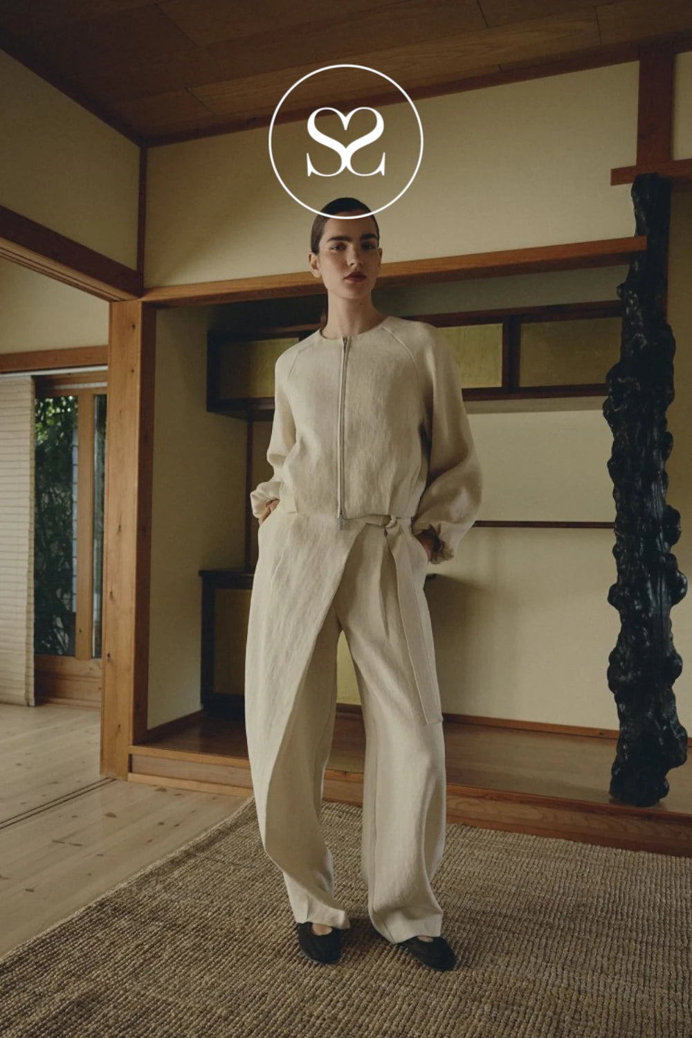 A model stands in a traditional Japanese room wearing a cream-colored textured zip-up jacket and matching wide-leg trousers. The image features a white circular logo overlay at the top center containing the stylized letters 'S2'.
