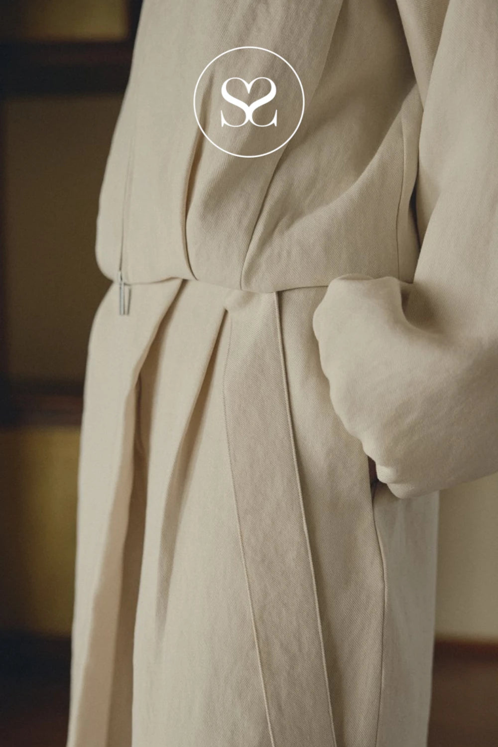 A close-up of a person wearing a light beige linen wrap or robe with a matching belt, featuring a white circular logo overlay with 'SC' at the top.