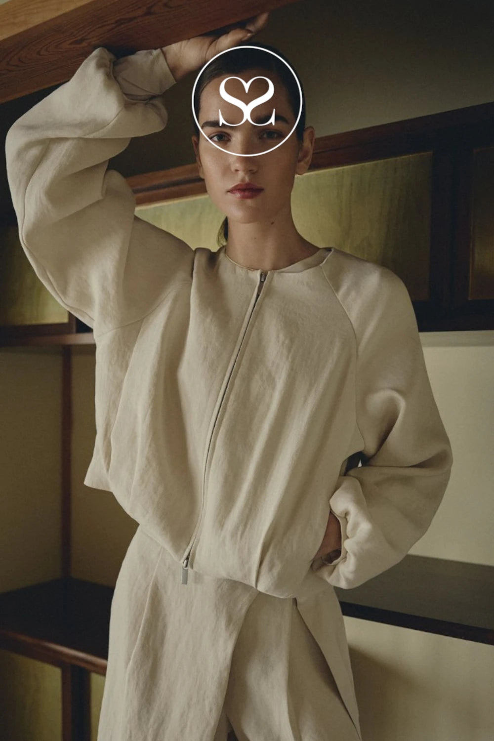 A model wearing a light beige, oversized zip-up jacket and matching pants in a warm-toned indoor setting. The model's face is obscured by a white circular logo overlay.